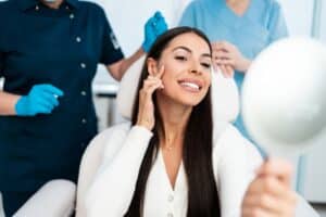 Woman sitting in a medical chair and smiling at her reflection in the mirror after a successful cosmetic procedure, such as microneedling with PRF, hyaluronic acid fillers, or botulinum toxin injections.