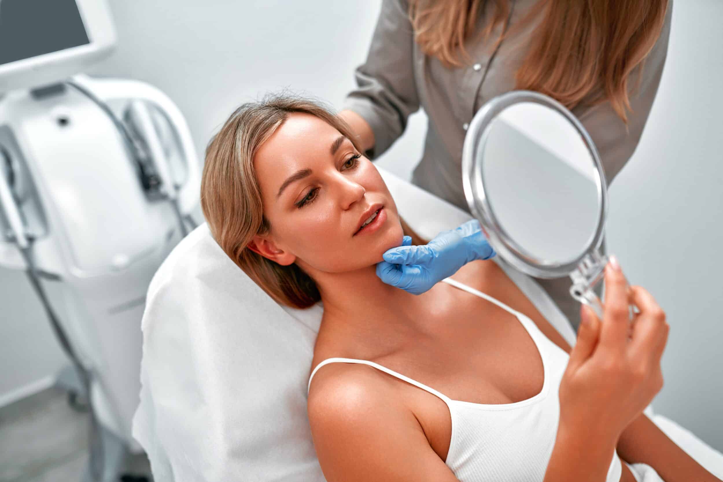Cosmetologist consulting a female client as she looks in the mirror at a beauty clinic, discussing PRF microneedling treatment and preparing for a skincare procedure in a spa setting