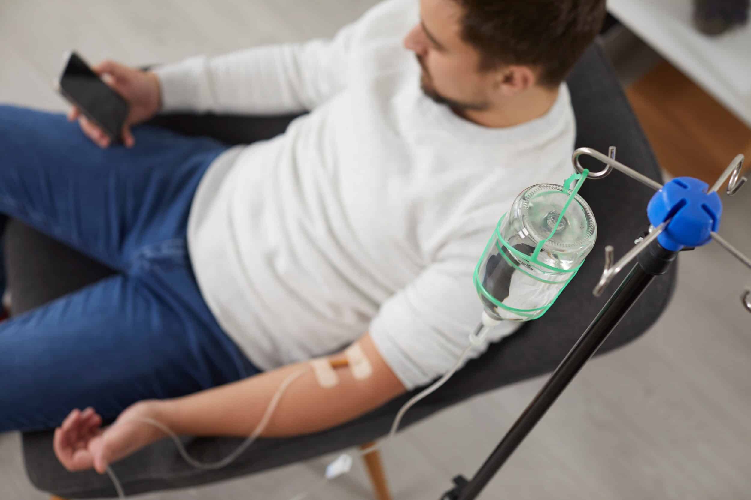 A man sits in a chair using his smartphone while receiving IV vitamin therapy through an infusion, highlighting modern wellness and injection treatment